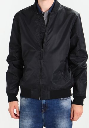 Man draagt een zwart bomberjack met rits over een grijs en marineblauw poloshirt en een blauwe spijkerbroek tegen een effen achtergrond.