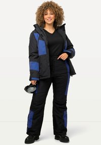 Zwarte winterjas met blauwe panelen, rits aan de voorkant en capuchon, gecombineerd met zwarte broek met blauwe accenten en dikke zolen.