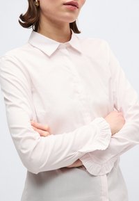 Γυναίκα με κοντά καστανά μαλλιά, φοράει χρυσούς κρίκους στα αυτιά και ανοιχτό ροζ μπλούζα με βολάν στα μανίκια, με σταυρωμένα τα χέρια, σε φωτεινό φόντο.