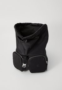 Zaino nero in nylon con parte superiore cinchata, due tasche anteriori con zip, accessori in argento e un logo impresso sulla tasca inferiore. Texture liscia.