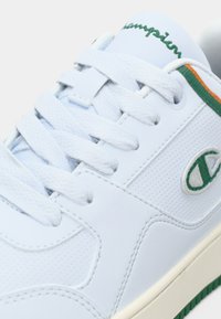 Λευκό αθλητικό παπούτσι με λείο συνθετικό επάνω μέρος, πράσινες και πορτοκαλί λεπτομέρειες, υφή στο πλάι, επίπεδα κορδόνια και πράσινη ραβδωτή λαστιχένια σόλα.