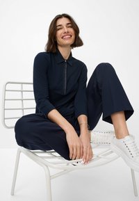 Marineblå langærmet polo med lynlåsdetalje, parret med løse marineblå bukser og hvide sneakers, siddende på en hvid metalstol.