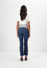Jean en denim bleu foncé avec une légère coupe bootcut, présentant un ourlet effiloché subtil et une étiquette en cuir brun marqué à la taille arrière.