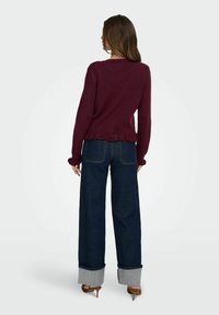 Pull en maille bordeaux avec un ourlet à volants, associé à un jean en denim foncé taille haute avec des poignets gris repliés et des chaussures à talons hauts.