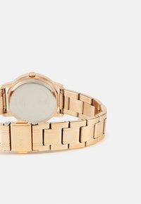 Rožinė auksinė laikrodžio, su metaline tinklelio apyranke, apvaliu korpusu ir minimalistiniu dizainu; turi lakuotą paviršių ir išgraviruotą prekės ženklą ant sagties.