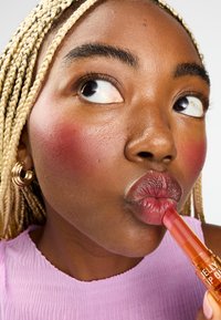 Huile à lèvres en gel dans un tube orange, tenue près de lèvres brillantes ; cheveux tressés ; portant un haut rose texturé ; maquillage naturel avec des joues rosées.
