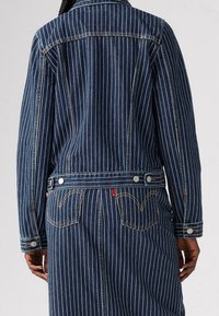 Farkkutakki, jossa pystysuorat valkoiset raidat, tummansininen väri, kaksi takataskua ompeludetaljeilla, metalliset napit ja hieman istuva malli.