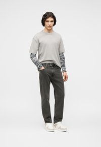 Νέος άνδρας όρθιος με τα χέρια στις τσέπες, φοράει πολλαπλές στρώσεις γκρι και μαύρα μακριά μανίκια με σχέδιο ζέβρας, μαύρα τζιν, ζώνη με καρφιά και λευκά αθλητικά παπούτσια.