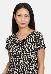 Zwarte en groene blouse met een zacht gewelfde halslijn, korte mouwen en een lichte stoftextuur, met abstracte vormen op een crèmekleurige achtergrond.