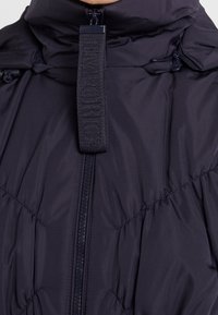 Marineblaue Steppjacke mit hohem Kragen, Frontreißverschluss und strukturiertem Riemen-Detail. Das Material wirkt weich und isoliert mit gestepptem Design.