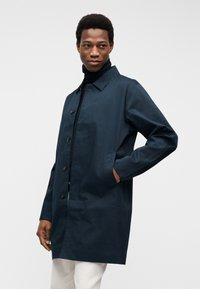 Άνδρας που φοράει μπλε σκούρο παλτό με κουμπιά και μαύρο ζιβάγκο, με το ένα χέρι στην τσέπη, κοιτάζοντας πλάγια σε απλό φωτεινό φόντο.