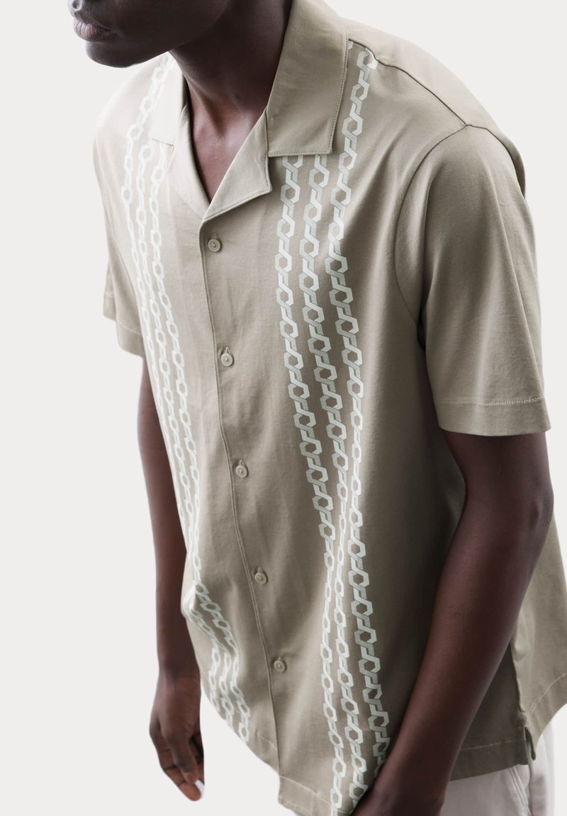 Camisa de manga corta beige con diseño de cuello, que presenta un patrón en cadena vertical en azul claro y un frente abotonado.