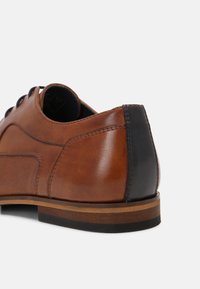 Chaussure en cuir marron avec une surface lisse, bout arrondi et accent de talon noir. Présente des panneaux latéraux texturés et un léger détail de couture.