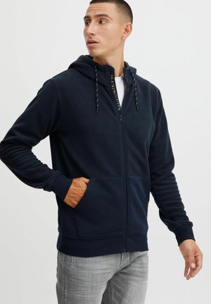 Sudadera con cremallera en color azul marino, fabricada en un tejido suave, con cordones amarillos, bolsillos delanteros y puños acanalados. La vista muestra a un modelo mirando hacia un lado.