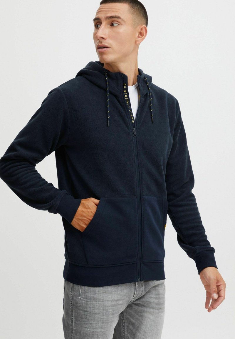 Sudadera con cremallera en color azul marino, fabricada en un tejido suave, con cordones amarillos, bolsillos delanteros y puños acanalados. La vista muestra a un modelo mirando hacia un lado.