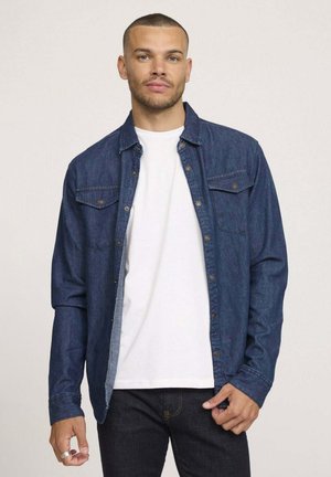 Chemise en jean foncé à boutons avec deux poches poitrine, manches longues et coupe décontractée. Portée par-dessus un t-shirt blanc uni et un jean foncé.