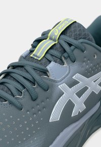 Grå atletisk sko med mesh-overdel, tekstureret overflade, kontrasterende hvidt logo og limegrøn accent på tungen og snørebåndene.