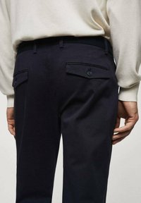 Pantalon marine en tissu à texture lisse, présentant deux poches arrière avec fermeture à bouton et une taille noire contrastante.
