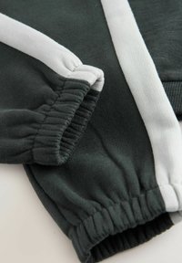 Dunkelgrüner Stoff mit weicher Textur, versehen mit weißen Streifen und elastischen Bündchen. Nahaufnahme, die die Nähdetails hervorhebt.