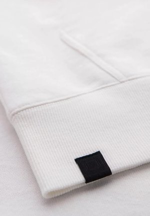 White cotton sweatshirt cuff with ribbed texture and a small black fabric label detail on the edge. Simple, clean design.