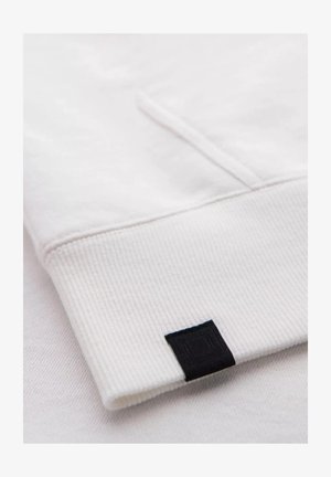 Poignet en coton blanc avec texture côtelée et un petit détail en étiquette noire sur le bord. Design simple et épuré.