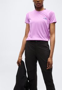 Άτομο που φοράει ροζ μπλουζάκι Patagonia και μαύρο παντελόνι, κρατώντας μια μαύρη ζακέτα μπροστά σε λευκό, απλό φόντο.