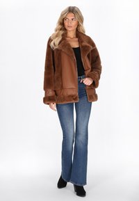 Vrouw met lang blond haar die een bruin nepbont jasje, een zwarte top, blauwe flared jeans en zwarte enkellaarsjes draagt, staand tegen een witte achtergrond.
