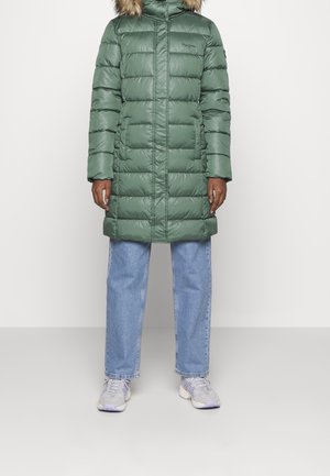 Persoon draagt een groene, gewatteerde lange winterjas met een capuchon met bontvoering, lichtblauwe spijkerbroek en witte sneakers, staande tegen een witte achtergrond.