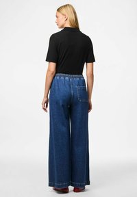 Platām kājām denimu bikses ar elastīgu jostu, tumši zilā krāsā, un aizmugurējo kabatu. Saskaņotas ar vienkāršu melnu īsām piedurknēm topu.