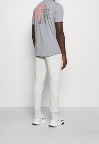 Hombre con una camiseta gráfica gris y pantalones jogger gris claro, con zapatillas deportivas blancas, de pie contra una pared blanca lisa.