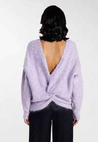 Lila, flauschiger Strickpullover mit tiefem V-Ausschnitt auf der Rückseite und gedrehter Gestaltung an der Taille. Lange Ärmel und lockere Passform. Kombiniert mit schwarzen Hosen.