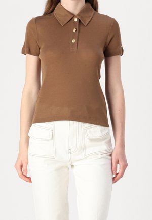 Femme portant un polo marron ajusté à manches courtes avec des boutons dorés, rentré dans un pantalon blanc avec de grandes poches frontales.