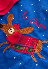 Tissu bleu à pois blancs avec un chien marron brodé portant une écharpe rouge à motif écossais, agrémenté d'un nœud rouge. Le texte indique « le Chien ».