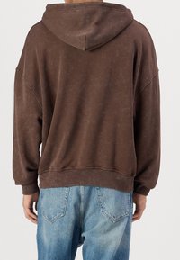 Sudadera marrón de gran tamaño con una textura desvanecida y una gran capucha, combinada con jeans de color azul claro. Presenta hombros caídos y un corte relajado.