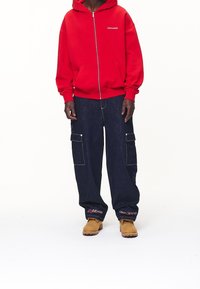 Person wearing red zip-up hoodie, dark blue cargo jeans with "Never Too Late" text near hems, and tan work boots standing on white background.