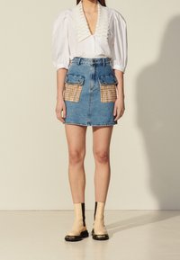 Blouse blanche avec un col détaillé, associée à une jupe en jean bleu avec des poches à carreaux. Le mannequin porte des bottines beige et noir.