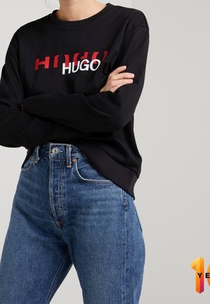 Person wearing black long-sleeve shirt with overlapping red and white "HUGO" logo, crossed arms, and high-waisted blue jeans against gray background.