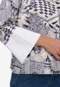 Top met witte manchetten, gemaakt van een blauw-wit patroonstof met speelse illustraties van gebouwen, bomen en geometrische vormen.