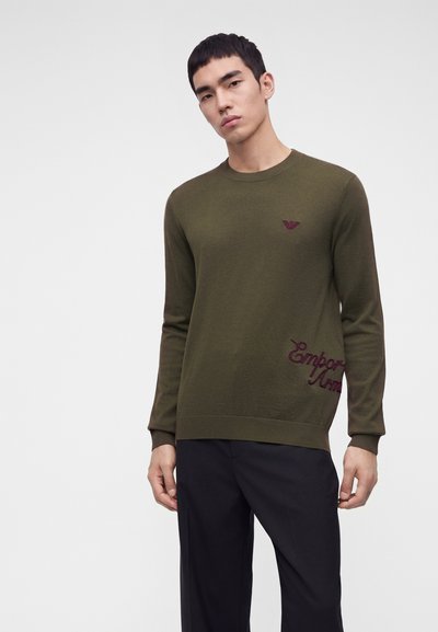 Homme portant un pull à manches longues vert olive avec un petit logo et un détail de texte, assorti à un pantalon noir, debout devant un fond clair uni.