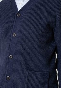 Close-up van een donkerblauwe gestructureerde gebreide cardigan met vier zwarte knopen en een voorzak over een lichtgestreepte shirt.