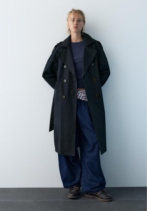 Femeie stă sprijinită de perete purtând un palton lung negru, cămașă bleumarin, pantaloni largi albastru închis, borsetă cu dungi și pantofi închiși la culoare.