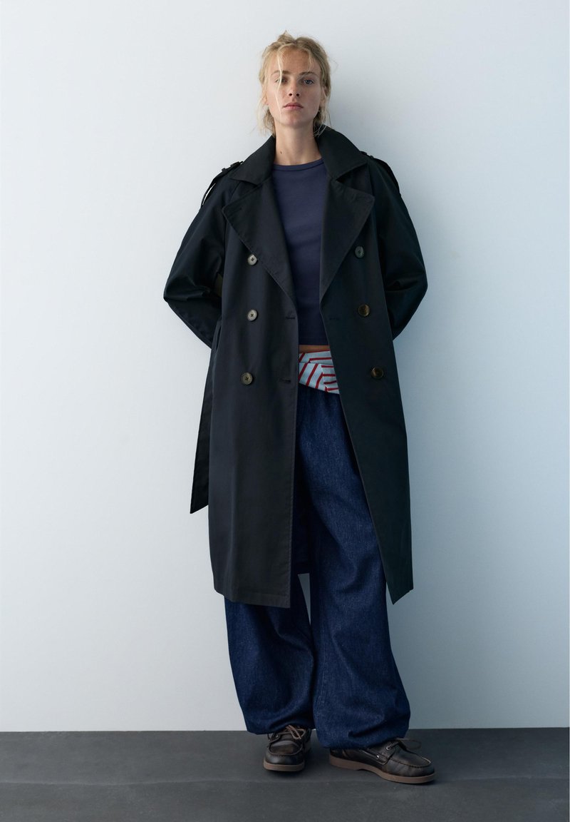 Donna in piedi contro il muro che indossa un lungo trench nero, camicia blu navy, pantaloni larghi blu scuro, marsupio a righe e scarpe scure.