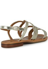 Sandal de tiras doradas metálicas con suela plana, que presenta múltiples tiras cruzadas y una correa con hebilla en el tobillo sobre una plantilla marrón.