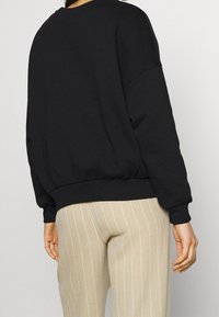 Felpa nera con scollatura rotonda, spalle scese e orlo arricciato. Abbinata a pantaloni beige a righe. Tessuto liscio.