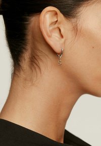 Pendiente de aro de plata con un charm colgante, que presenta una letra 'S' estilizada adornada con pequeñas piedras, colgando de una superficie lisa.