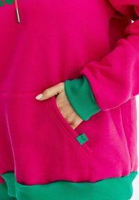 Różowa bluza z zielonym ściągaczem u dołu i na mankietach. Z przodu kieszeń z małą zieloną metką. Gładka tekstura materiału.