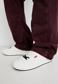 Sneakers bianchi con lacci neri, dotati di dettagli perforati e un logo rosso, abbinati a pantaloni a sigaretta bordeaux. Design pulito e texture liscia.