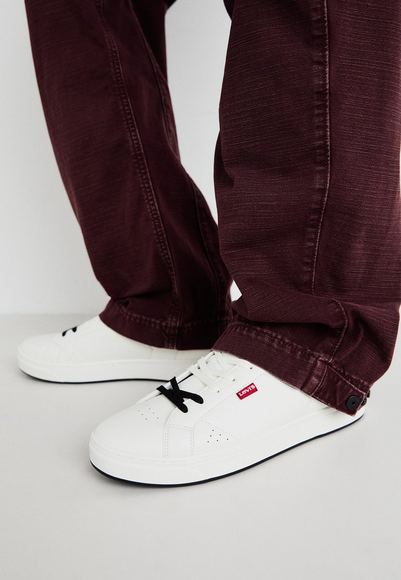 Sneakers bianchi con lacci neri, dotati di dettagli perforati e un logo rosso, abbinati a pantaloni a sigaretta bordeaux. Design pulito e texture liscia.