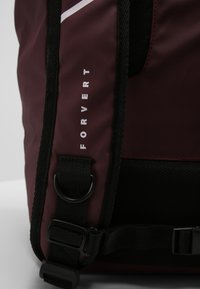Sac à dos bordeaux avec des accents noirs, fabriqué en tissu durable, avec des bretelles ajustables, et le mot "FORVERT" imprimé verticalement en blanc.