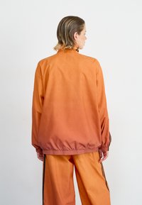 Oransje gradientjakke med høy krage og elastisk hem, kombinert med matchende bukser med svarte sidestriper. Glatt tekstur.